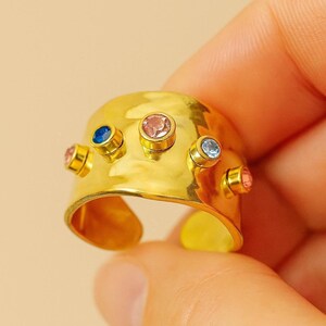 Peut inclure: Une bague dorée à large bande et design ouvert. La bague est ornée de cinq pierres précieuses colorées : rose, bleu et bleu clair. Les pierres sont serties dans de petits chatons dorés.