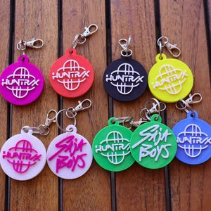 May include: A collection of colorful keychains with silver clasps. The keychains are round with the text "HUNTRA" and "Skit Boys" in white. Colors include pink, red, black, yellow, white, green, and blue. The keychains are on a wooden surface.