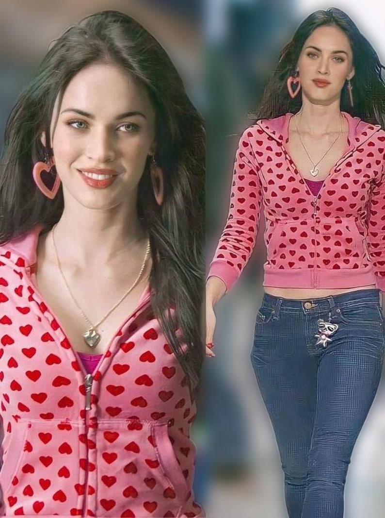 May include: A pink hooded top with red heart motifs, worn with blue jeans. The model is also wearing heart-shaped earrings and a necklace with a heart pendant. The outfit is a casual, romantic style.