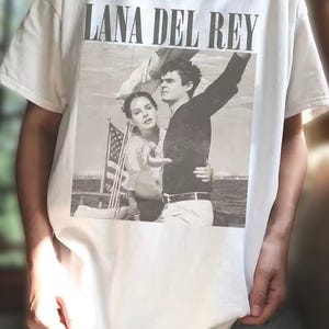 Puede incluir: Camiseta blanca con una imagen en blanco y negro de Lana Del Rey y un hombre. El texto "LANA DEL REY" est&aacute; impreso en letras negras en la parte superior de la imagen. La imagen muestra a la pareja en un barco con una bandera estadounidense.