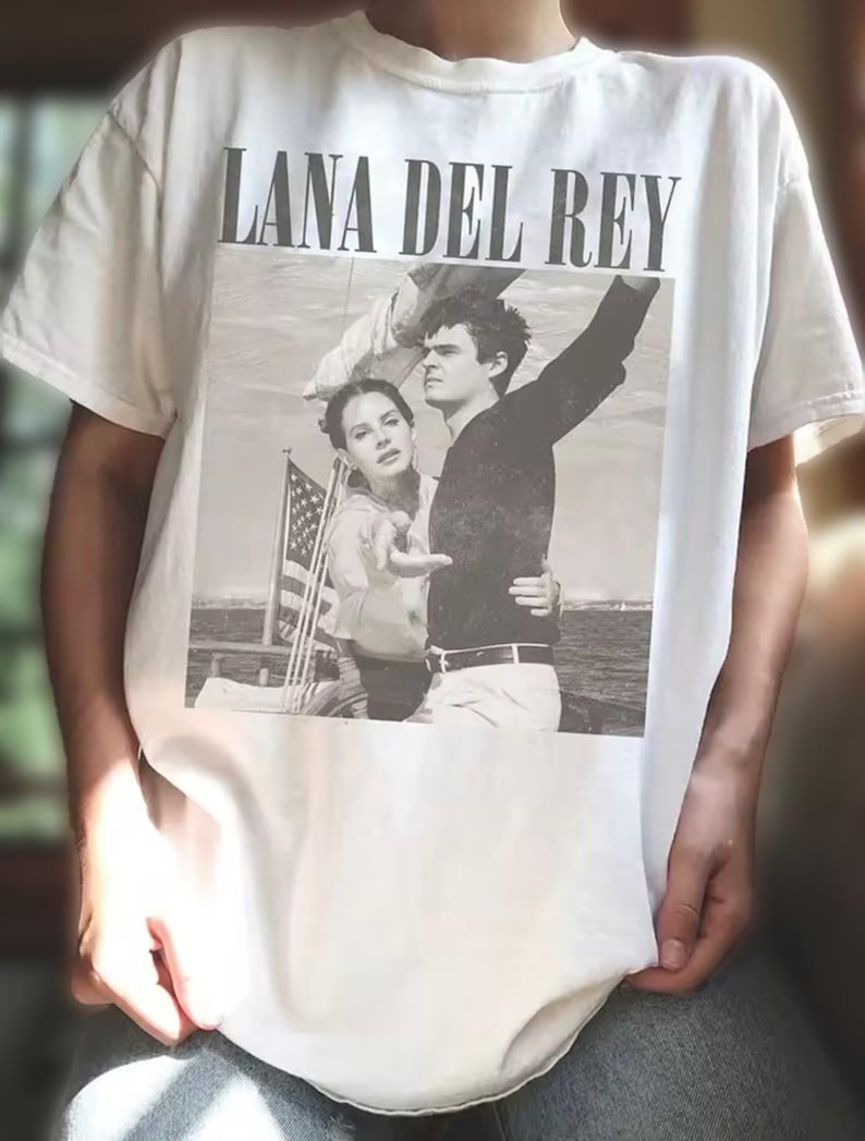 Puede incluir: Camiseta blanca con una imagen en blanco y negro de Lana Del Rey y un hombre. El texto "LANA DEL REY" est&aacute; impreso en letras negras en la parte superior de la imagen. La imagen muestra a la pareja en un barco con una bandera estadounidense.