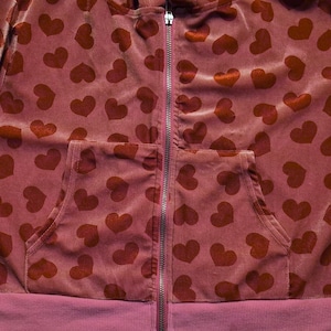 May include: A burgundy zip-up hoodie featuring a pattern of dark red hearts. The hoodie has a front pocket and ribbed hem and cuffs. The zipper is silver.