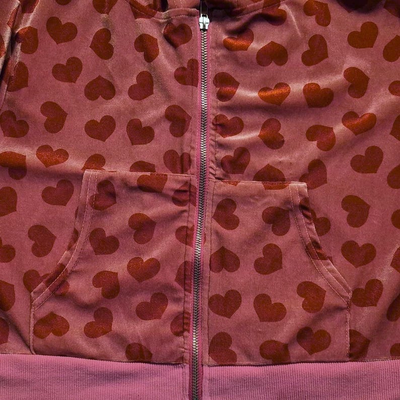 May include: A burgundy zip-up hoodie featuring a pattern of dark red hearts. The hoodie has a front pocket and ribbed hem and cuffs. The zipper is silver.