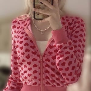 May include: Pink zip-up hoodie with a red heart pattern. The jacket has a cropped fit, long sleeves, and a ribbed hem and cuffs. A white top is visible underneath. A pearl necklace is worn.