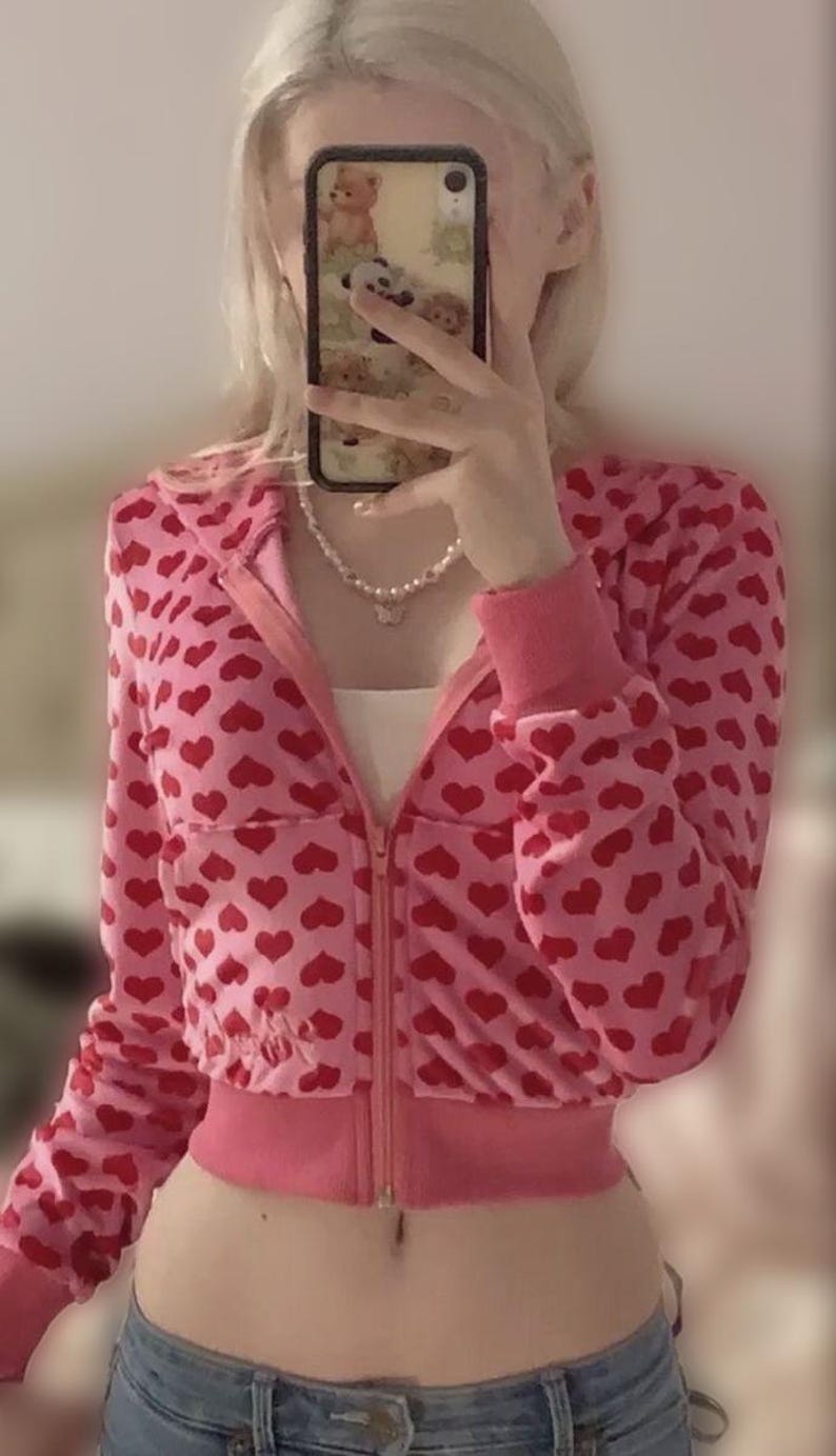 May include: Pink zip-up hoodie with a red heart pattern. The jacket has a cropped fit, long sleeves, and a ribbed hem and cuffs. A white top is visible underneath. A pearl necklace is worn.