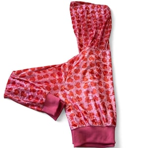May include: A pink hooded sweatshirt with a pattern of red hearts. The cuffs and hem are a darker shade of pink. The fabric has a soft, velvety texture.