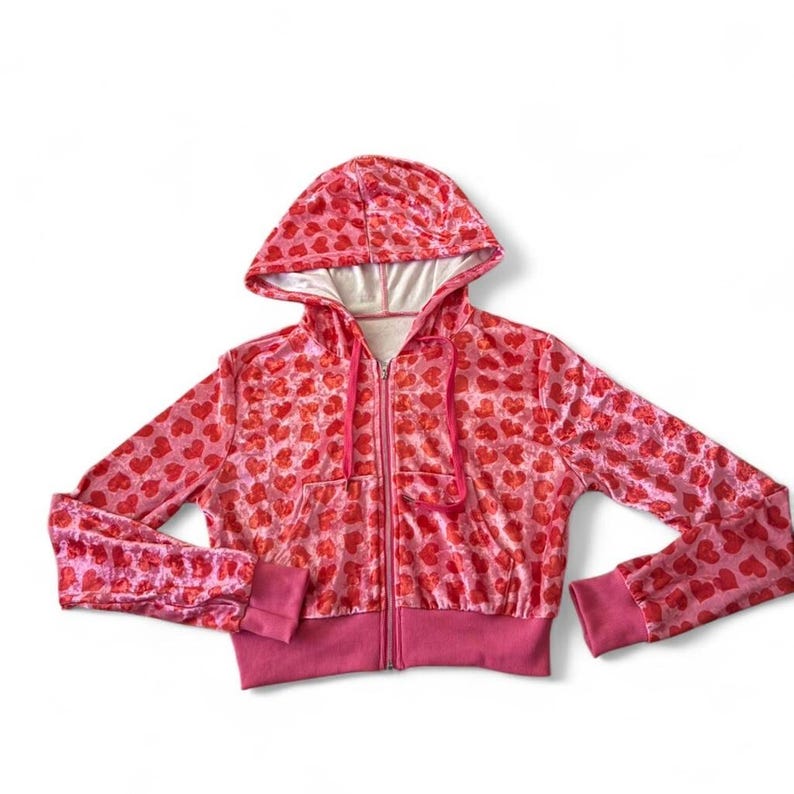 May include: A pink zip-up hoodie with a heart pattern. The hoodie has a drawstring hood, a full-length zip, and ribbed cuffs and hem. The heart pattern is in a darker shade of red, and the hood's interior is white.