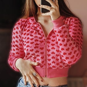 May include: A pink cropped zip-up jacket with a red heart pattern. The jacket has long sleeves and a ribbed hem and cuffs. The model is wearing blue jeans.