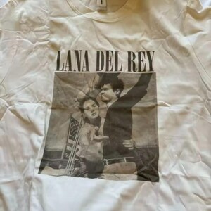 Puede incluir: Camiseta color crema con el texto "LANA DEL REY" sobre una imagen en blanco y negro de dos personas. La imagen parece ser una fotograf&iacute;a. La camiseta est&aacute; hecha de un material suave.