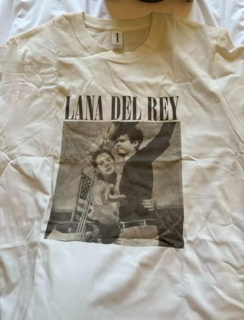 Puede incluir: Camiseta color crema con el texto "LANA DEL REY" sobre una imagen en blanco y negro de dos personas. La imagen parece ser una fotograf&iacute;a. La camiseta est&aacute; hecha de un material suave.