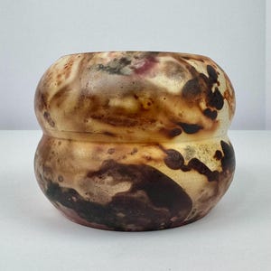 May include: A handcrafted ceramic vessel with a unique, organic shape. The exterior features a blend of warm colors, including browns, tans, and hints of burgundy, creating a marbled effect. The vessel has a bulbous form with a constricted middle section.