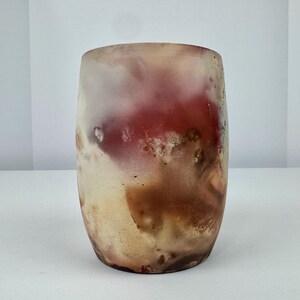 May include: A cylindrical, handcrafted glass vase with a unique, abstract design. The vase features a blend of colors, including red, brown, and cream, creating a marbled effect. The glass has a slightly frosted appearance.