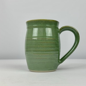 May include: A handmade green ceramic mug with a curved handle. The mug has a cylindrical shape with horizontal lines and a slightly flared rim. The interior and handle are the same shade of green, with a lighter green rim.