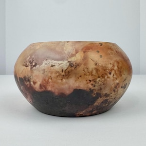 May include: A round, handcrafted ceramic bowl with a unique, organic design. The bowl features a blend of earthy tones, including shades of brown, beige, and reddish-brown, with a dark brown base. The surface has a textured appearance.