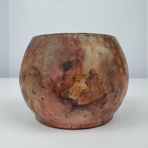 May include: A round, ceramic vessel with a wide opening. The exterior features a mottled pattern of brown, tan, and reddish-orange hues, creating a textured appearance. The vessel has a slightly rounded base and a smooth rim.