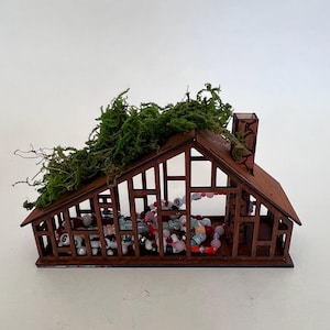 May include: A miniature wooden house with a brown roof and walls, filled with colorful beads and topped with green moss. The house has a chimney and open sides, creating a decorative display.