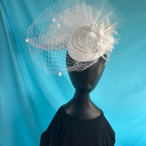 Puede incluir: Un sombrero fascinador blanco con velo, perlas y adornos florales. El sombrero presenta una pequeña base redonda adornada con una banda ribeteada de perlas y un grupo de flores de tela blanca. Un delicado velo con adornos de perlas cubre el sombrero.