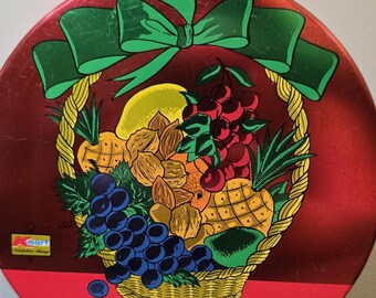 May include: A round, red tin with a decorative illustration of a fruit basket and a green bow. The basket contains various fruits, including grapes, cherries, pineapple, and a lemon. The text "Fruit Cake" is written in cursive below the basket.