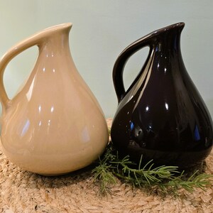 May include: Two ceramic pitchers, one beige and one dark brown, with curved handles. The pitchers have a teardrop shape and sit on a woven surface. Green sprigs are placed between the pitchers.