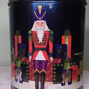 May include: A cylindrical, dark blue tin with a decorative image of a nutcracker soldier. The nutcracker is dressed in a red jacket, purple pants, and a tall hat. The tin is decorated with gift boxes.