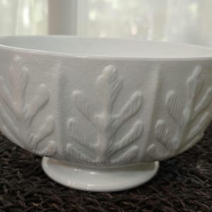 May include: A white ceramic bowl with a pedestal base. The bowl features a repeating leaf pattern around the exterior. The bowl is approximately 8 inches in diameter and 4 inches tall.