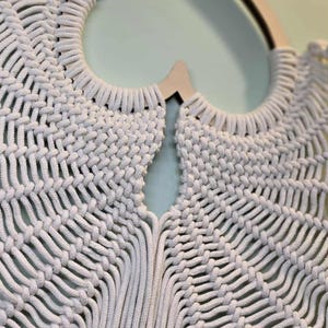 May include: Close-up of a cream-colored macrame wall hanging. The intricate design features woven knots and a heart-shaped wooden frame. The texture and pattern create a bohemian aesthetic, perfect for home decor.