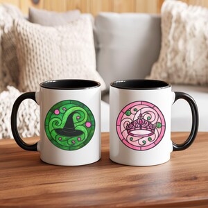Pu&ograve; includere: Due tazze in ceramica bianca con manici e bordi neri. Una tazza presenta un design a cappello da strega verde e nero, l'altra una corona rosa. Entrambi i disegni sono in uno stile circolare a vetrata.