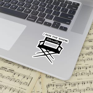 May include: A black and white sticker featuring a director's chair with the text "I RUN THE SHOW, literally." The sticker is placed on a laptop, with sheet music visible in the background.