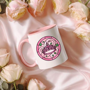Pu&ograve; includere: Tazza in ceramica bianca con interno e manico rosa. La tazza presenta un disegno circolare di una corona rosa con accenti verdi, simile a una vetrata. La tazza &egrave; circondata da rose rosa e poggia su una superficie di raso rosa.