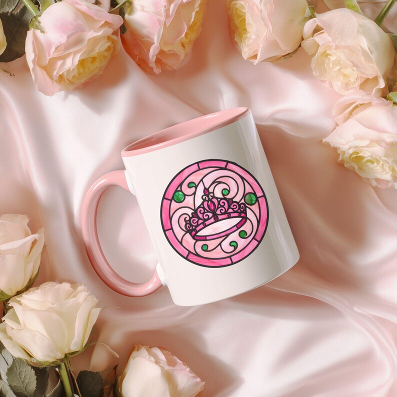 Pu&ograve; includere: Tazza in ceramica bianca con interno e manico rosa. La tazza presenta un disegno circolare di una corona rosa con accenti verdi, simile a una vetrata. La tazza &egrave; circondata da rose rosa e poggia su una superficie di raso rosa.