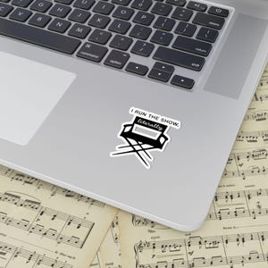 May include: A black and white sticker featuring a director's chair with the text "I RUN THE SHOW, literally." The sticker is placed on a laptop, with sheet music visible in the background. The sticker is a fun and playful item.