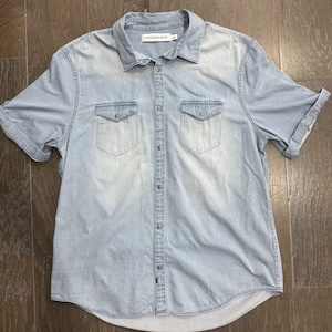 Könnte beinhalten: Hellblaues Jeanshemd mit kurzen Ärmeln, zwei Brusttaschen und Knopfleiste. Das Hemd hat einen klassischen Kragen und das Calvin Klein Jeans-Logo ist auf dem Etikett sichtbar.