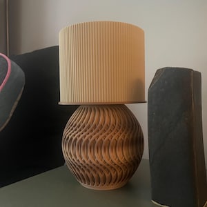 Puede incluir: Una lámpara de mesa decorativa con una pantalla cilíndrica beige y una base esférica de madera con un diseño tallado en forma de ola. Un objeto oscuro y rectangular está al lado de la lámpara.