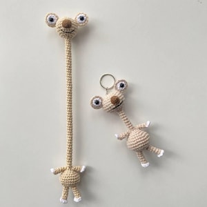 May include: Two beige crochet alien figures. One is a long, slender figure with a small body and outstretched arms. The other is a keychain version with a round body and a keyring. Both have large eyes, a brown nose, and white accents.