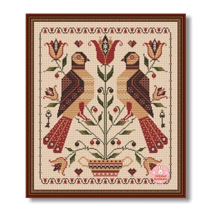 May include: Framed cross-stitch artwork featuring two stylized birds flanking a central floral design. The birds have brown and tan bodies with red and black accents. The artwork is set against a beige background and framed in a dark brown wooden frame.