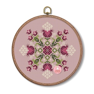 May include: A cross-stitch embroidery with a floral pattern in pink, green, and white on a light pink backdrop. It's displayed in a round wooden hoop with a brass hanger. The text "Stitched Sunbeams" is visible on the lower right of the embroidery.