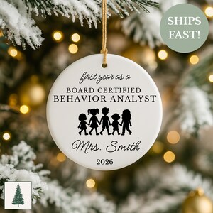 Peut inclure: Ornement de Noël rond en céramique blanche avec une ficelle dorée. Il présente le texte "first year as a BOARD CERTIFIED BEHAVIOR ANALYST", une silhouette de cinq enfants, le nom "Mrs. Smith" et l'année "2026". Décoration de vacances.