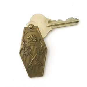 以下が含まれることがあります： 棺桶の形をした真鍮色のキーホルダーが付いた金属製の鍵。キーホルダーの表面には、上部に貝殻、装飾的なスクロール模様、そして縦縞の背景の上に交差した2本の鍵がエンボス加工でデザインされています。