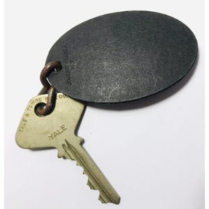 May include: A vintage key ring with a dark gray oval tag and a Yale key. The tag is embossed with "JOHNNIE WALKER N.Y." and the key has "YALE & TOWNE CO." and "YALE" printed on it. The key ring is made of metal.
