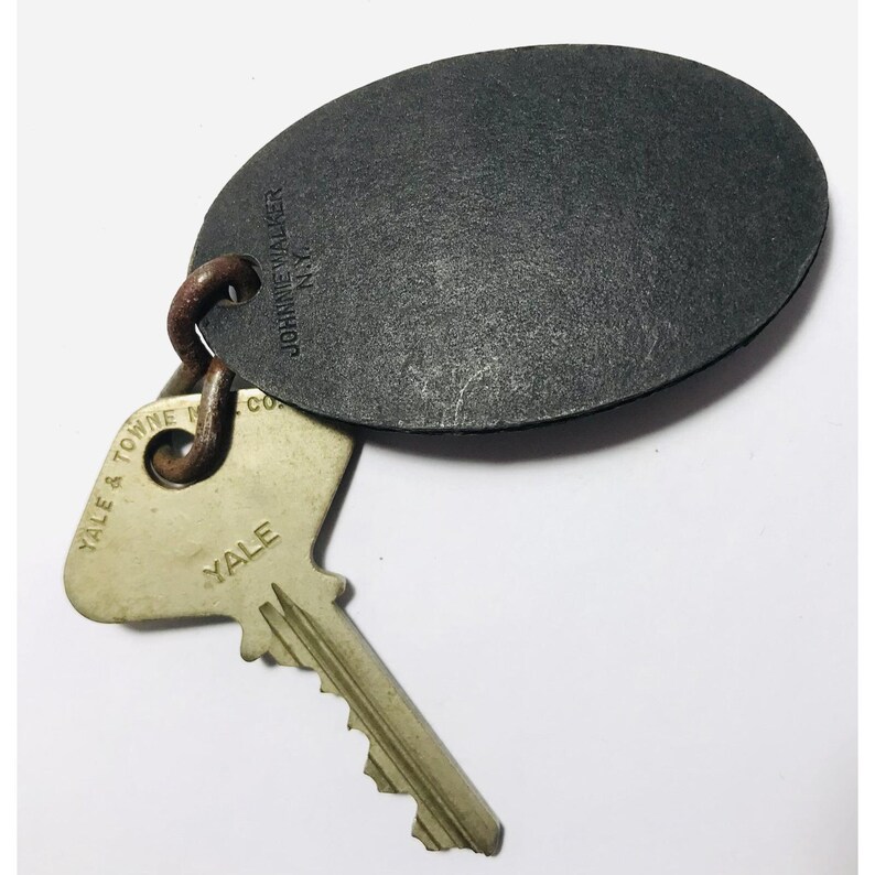 May include: A vintage key ring with a dark gray oval tag and a Yale key. The tag is embossed with "JOHNNIE WALKER N.Y." and the key has "YALE & TOWNE CO." and "YALE" printed on it. The key ring is made of metal.