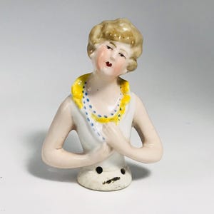 Könnte beinhalten: Vintage Porzellan-Puppen-Halbfigur mit weißem Oberteil und gelbem Kragen mit blauen Tupfen. Die Puppe hat blondes Haar, rosige Wangen und rote Lippen. Die Figur ist eine Büste, die Arme sind vor der Brust verschränkt.