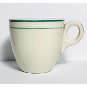 May include: Cream-colored ceramic coffee cup with a rounded body and a handle. The cup features two thin, parallel green stripes near the rim. The cup is set against a white background.