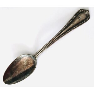 May include: A vintage silver spoon with an oval bowl and a long handle. The handle has a decorative design and a monogrammed initial "M" at the top. The spoon shows signs of age and wear, with a tarnished appearance.