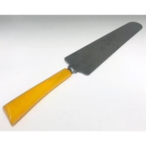 May include: A cake server featuring a long, rectangular, dark grey blade and a bright yellow handle. The handle is smooth and rounded, and the blade is attached with a silver metal piece. The server is designed for cutting and serving cake.