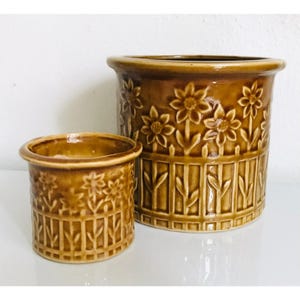 May include: Two brown ceramic flower pots. The larger pot has a cylindrical shape with a wide rim and is decorated with embossed flowers and vertical lines. The smaller pot has a similar design. Both pots are on a white surface.
