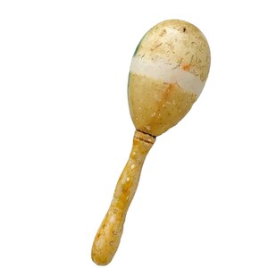 Puede incluir: Una maraca vintage con una cabeza en forma de huevo marr&oacute;n claro y un mango largo de madera. La cabeza presenta una banda blanca y detalles verdes. El mango es de un tono m&aacute;s claro con peque&ntilde;os puntos oscuros.