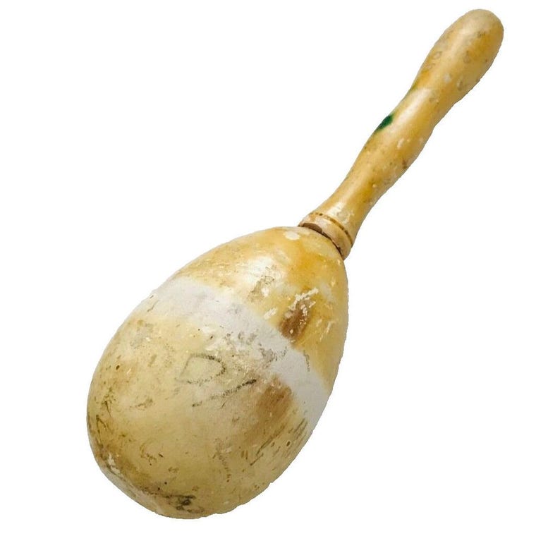 Puede incluir: Una maraca de madera vintage con un mango marr&oacute;n claro y un cuerpo en forma de huevo. El cuerpo tiene una banda blanca y muestra signos de desgaste. La maraca es un instrumento musical, probablemente utilizado para el ritmo y la percusi&oacute;n.