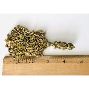 May include: A gold-toned metal decorative piece with an ornate design. The piece is shaped like a fan and has a long handle. It measures approximately 5 cm in length.