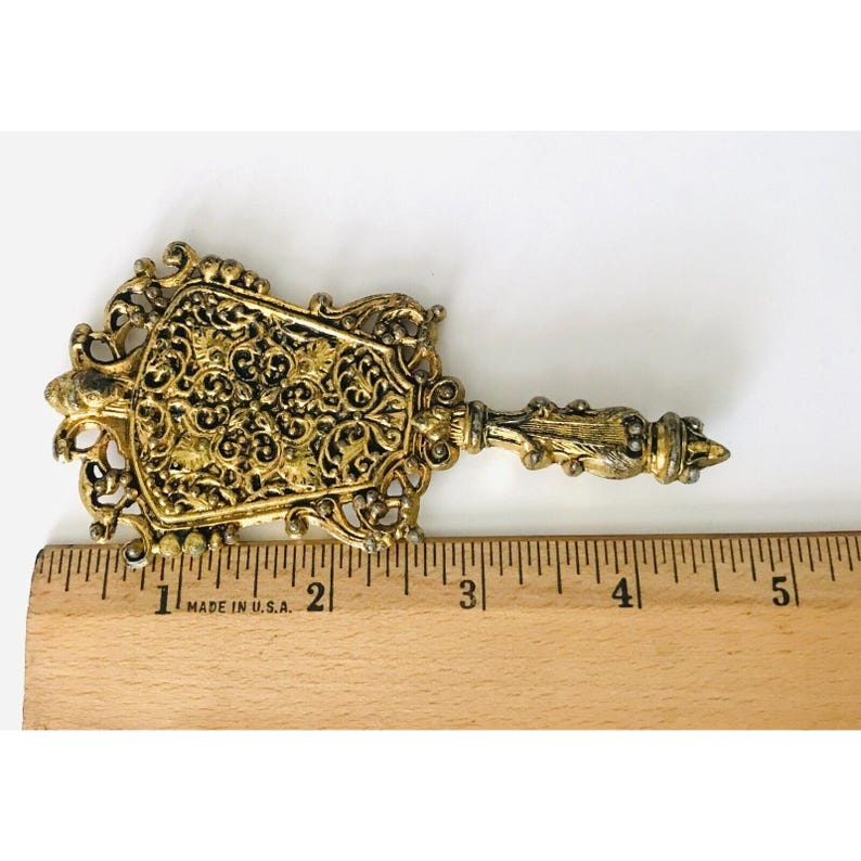 May include: A gold-toned metal decorative piece with an ornate design. The piece is shaped like a fan and has a long handle. It measures approximately 5 cm in length.