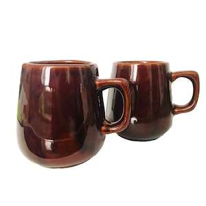 May include: Two brown ceramic mugs with a glossy finish. Each mug has a rounded body and a square handle. The mugs are a deep, rich brown color and appear to be handmade. The mugs are set against a white background.